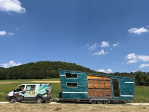 A turquoise wooden tiny house on a trailer being towed by a white van in a rural field with a tree-covered hill in the background.