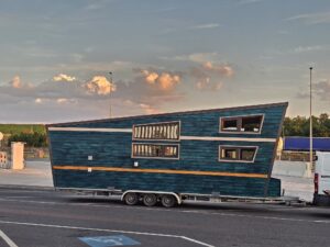 Tilted blue wooden building mounted on a trailer in a parking lot with cloudy sky and tall street lamps nearby.