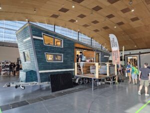 Teal wooden tiny house on a trailer displayed inside a convention center, with visitors inspecting it and banners nearby.